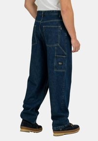 Pantalones de mezclilla azul oscuro de corte holgado, con múltiples bolsillos grandes, costuras visibles y un parche con el logo en el bolsillo trasero.