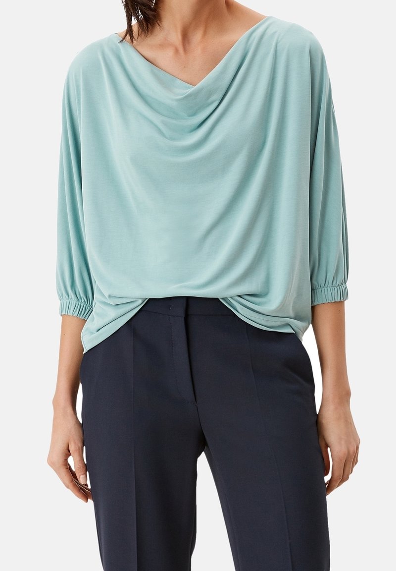Top drappeggiato di colore azzurro chiaro con maniche a 3/4 e polsini arricciati, abbinato a pantaloni scuri. Il tessuto sembra morbido e liscio.