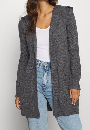 Cardigan en tricot gris avec capuche, doté de deux poches avant et de poignets côtelés, associé à un haut blanc et un jean bleu clair.
