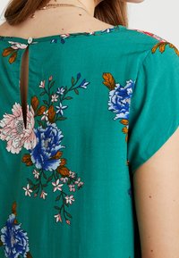 Květinová halenka v tyrkysovém materiálu s růžovými, modrými a oranžovými květinovými vzory, s uzávěrem na knoflík na zadní straně a krátkými rukávy.