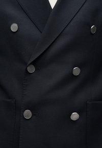 Κοντινό πλάνο ενός διπλής σειράς μπλέ σκούρου σακακιού με έξι στρογγυλά μεταλλικά κουμπιά από βουρτσισμένο ασημί και υφασμάτινο ύφος με υφή.