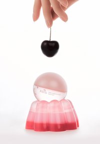 Masque pour les lèvres au collagène dans un contenant sphérique rose posé sur une base gélatineuse rose ; une cerise noire est suspendue au-dessus.