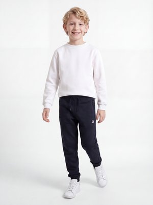 Jeune garçon souriant aux cheveux blonds, portant un sweat blanc, un pantalon de survêtement noir et des baskets blanches, marchant vers l'avant sur un fond blanc.