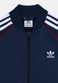 Marineblaues Zip-Jackett mit geripptem Kragen, das drei weiße Streifen auf den Schultern, rote Akzente und ein kleines weißes Adidas-Logo auf der Brust aufweist.