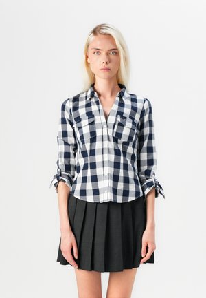 Navy and white checkered button-up shirt with roll-up sleeves and two front pockets, paired with a pleated black skirt.