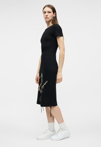 Γυναίκα που στέκεται φορώντας μαύρο midi φόρεμα με κοντά μανίκια και μπεζ γράμματα στη μία πλευρά, φοράει λευκές κάλτσες και λευκά αθλητικά παπούτσια, σε λευκό φόντο.