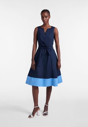 Donna in piedi che indossa un abito senza maniche blu scuro con orlo azzurro chiaro e vita annodata, abbinato a sandali bianchi con tacco alto su uno sfondo bianco uniforme.