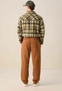 Homme portant une veste à carreaux verte et marron, un pantalon en velours côtelé marron, des baskets blanches et une casquette grise, se tenant les mains dans les poches, de dos.