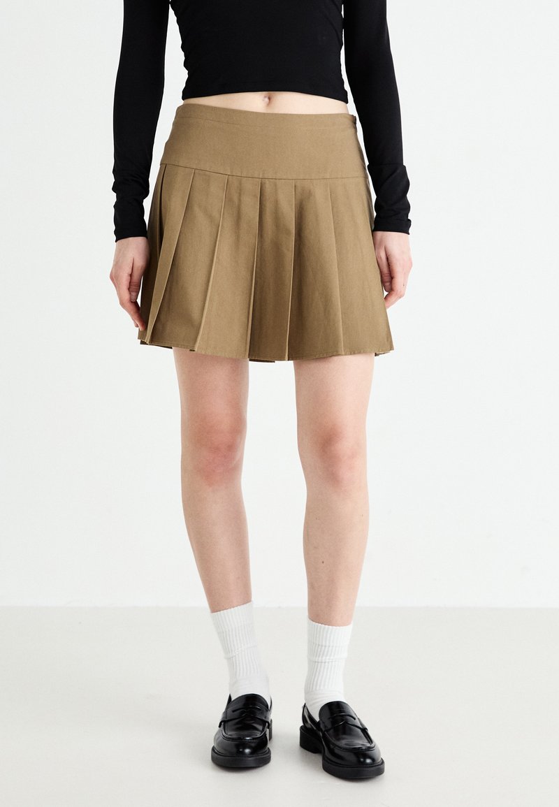 Jupe mini plissée kaki avec une taille ajustée, présentant des plis marqués et une texture lisse. Associée à des mocassins noirs et des chaussettes blanches.