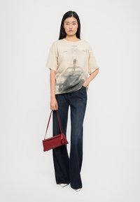 Camiseta oversized beige con estampado gráfico, combinada con jeans acampanados de color azul oscuro. El modelo sostiene un bolso de cuero rojo y lleva tacones blancos.