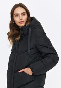 Czarna kurtka puchowa z kapturem, o wzorze pikowanym, z elastycznymi mankietami i kieszeniami, wykonana z gładkiego, lekko błyszczącego materiału.