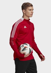 Rotes, langärmliges Sportshirt mit weißem Kragen und drei weißen Streifen an den Ärmeln, kombiniert mit einer schwarzen Hose und einem weißen Fußball mit bunten Akzenten.