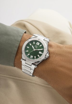 Silberne Metallarmbanduhr mit grünem Zifferblatt, römischen Zahlen, Datumsanzeige und drei Zeigern, getragen am linken Handgelenk mit grünem Hemdärmel.