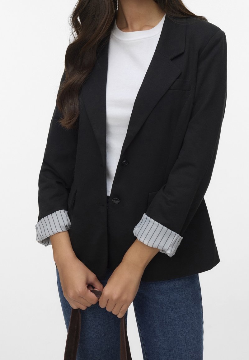 Femme portant un blazer noir avec des poignets rayés roulés, une chemise blanche et un jean bleu, tenant des poignées de sac marron devant elle.