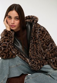 Giovane donna con lunghi capelli scuri, indossa una giacca in pelliccia sintetica marrone con stampa leopardata, un top marrone e jeans blu, mentre poggia il viso su una mano.