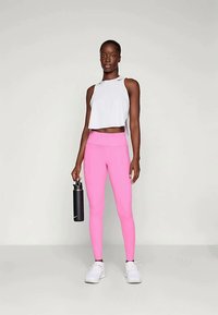 Sportos öltözet, amely egy cropped fehér topot és egy testhezálló rózsaszín leggingset tartalmaz. Egy vízpalack van a kezében; a fehér sportcipők teszik teljessé a megjelenést.