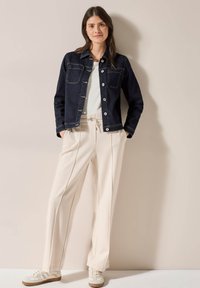 Veste en denim foncé avec deux poches poitrines, boutons blancs et détails de couture. Assortie à un pantalon plissé couleur crème et des baskets claires.
