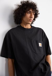 Young person with curly hair and septum piercing wearing a black Carhartt T-shirt with a front pocket, leaning against a white wall.