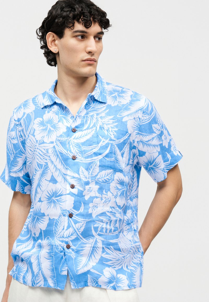Man met krullend haar, gekleed in een blauw shirt met korte mouwen en een witte bloemprint, kijkt naar links tegen een effen achtergrond.
