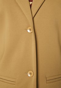 Gros plan sur un blazer beige montrant deux boutons beiges, des boutonnières, des revers et des poches horizontales fendues cousues à l'avant.