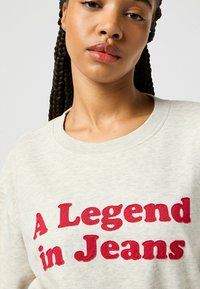 Felpa grigia chiaro con testo ricamato rosso in bold "A Legend in Jeans", con colletto a giro e un tessuto morbido e textured.