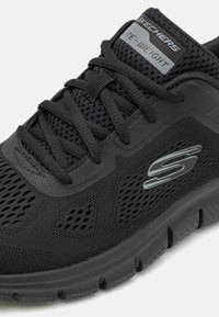 Black athletic sneaker with a mesh upper, rounded toe, and textured sole. Features laces and a logo on the side and tongue.