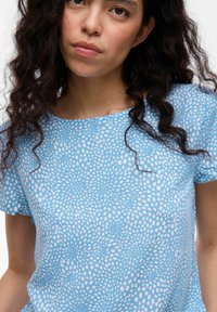 Ljusblå blus med kort ärm som har ett prickigt mönster i vitt, med en avslappnad passform och mjuk tygstruktur.