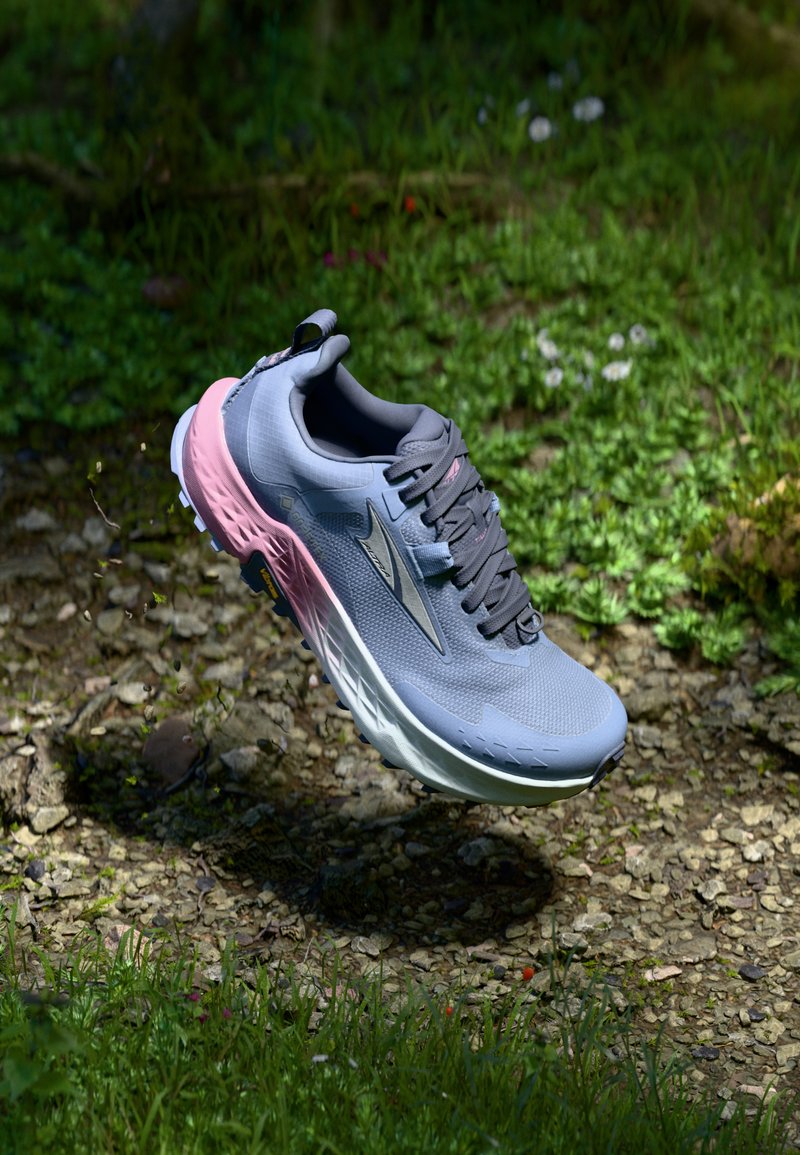 Lightweight running shoe in gray with pink accents, textured upper, cushioned sole, and flat laces, suspended above a natural stone and grass surface.