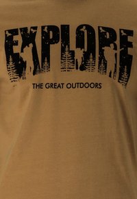Ruskea puuvillainen t-paita, jossa on suuri musta graafinen kuva, joka sanoo "EXPLOROITSE", ja siinä on puiden ja vaeltajien siluetteja. Teksti alapuolella lukee "SUURI LUONTO".
