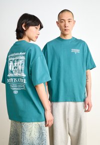 Teal cotton t-shirts feature graphic prints on the back and front. The female model wears a patterned skirt, while the male model wears gray sweatpants.