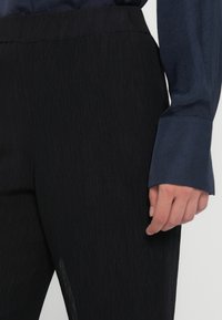 Zwarte, gestructureerde broek met een slanke pasvorm. Een donkerg blauw, glad shirt is zichtbaar, met een knoopmanchet en een getailleerde kraag.