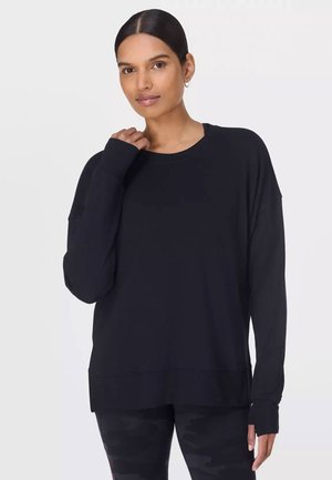 Vrouw met donker haar in een lage knot, gekleed in een oversized, lange mouwen zwarte trui en zwarte camouflagelegging, staand tegen een egale achtergrond.