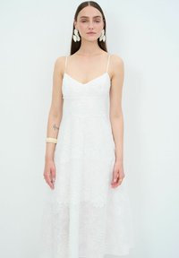 Witte jurk met dunne spaghettibandjes, bloemenborduursel, vloeiende rok en een gelaagd ontwerp. Model draagt crèmekleurige oorbellen en een armband.