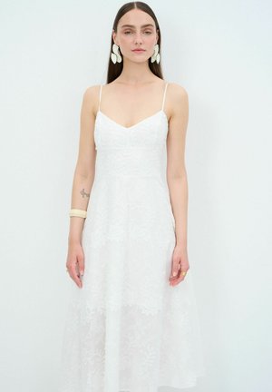 Abito bianco con spalline sottili, ricami floreali, gonna fluida e design a strati. La modella indossa orecchini color crema e un braccialetto.