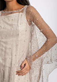 Vestido de un beige claro con una capa externa de tejido abierto y texturizado. Presenta un escote cuadrado y mangas largas y transparentes. Textura de tela suave.