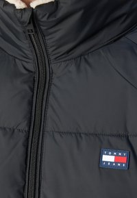 Schwarze gesteppte Jacke mit Vorderreißverschluss und einem Tommy Jeans Logo-Patch auf der Brust, mit einem weißen und roten Streifendetail.