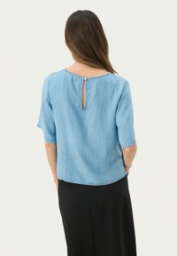 Lysblå denimbluse med rund halsudskæring, halvlange ærmer og nøglehulslukning bagpå, der har en knaplukning. Glat tekstur, afslappet pasform.