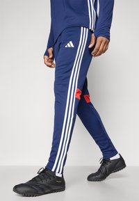 Marineblauwe sportbroek met witte strepen en rode accenten, met een taps toelopende pasvorm en een elastische tailleband. Gedragen met zwarte sportschoenen.