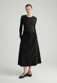 Kobieta stojąca z rękami w kieszeniach, ubrana w czarną bluzkę z długim rękawem, zapinaną na guziki czarną midi spódnicę oraz czarne buty typu Mary Jane na szarym tle.