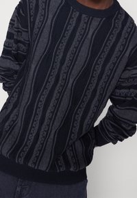 Tmavý pletený svetr s texturovanými svislými pruhy v různých tmavých odstínech šedé. Má žebrovaný výstřih a manžety.