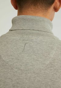 Le pull en maille grise présente un col montant, une texture côtelée et un yoke circulaire avec un accent brodé au dos.