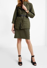 Veste en tissu vert olive avec quatre poches et une fermeture éclair, portée sur une jupe assortie. Une ceinture noire souligne la taille ; sandales à talons hauts noires.