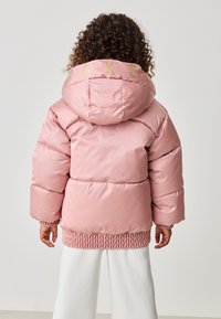 Lyserød boblejakke med hætte, der har ribbetrukne kanter ved bunden og ærmerne. Bogstaverne "BAKE" i guld på bagsiden. Glat, skinnende stofoverflade.