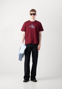 Bordo pamučna majica s logom, crne široke traperice i crne cipele. Svijetloplava jakna prebačena preko jednog ramena. Crne sunčane naočale.