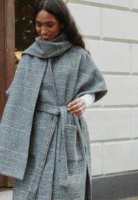 Hall triibuline rühtmantel tekstuurse kangaga, kõrge krae ja laiade varrukatega, vöökoha juures lipsuga detailiga.