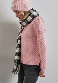 Ljusrosa stickad tröja med strukturerat mönster, tillsammans med en svart och vit rutad halsduk med fransar och mörka jeans.