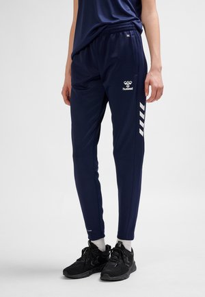 Osoba ubrana w granatowe spodnie dresowe marki Hummel z elastycznym pasem i czarne buty sportowe stojąca na jednolitym tle.