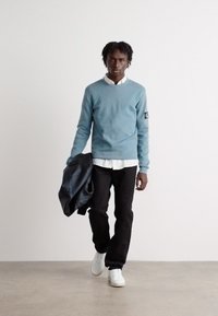 Lys blå genser med teksturert stoff, lagdelt over en hvit skjorte. Sorte bukser og hvite sneakers. Holder en svart skinnjakke.