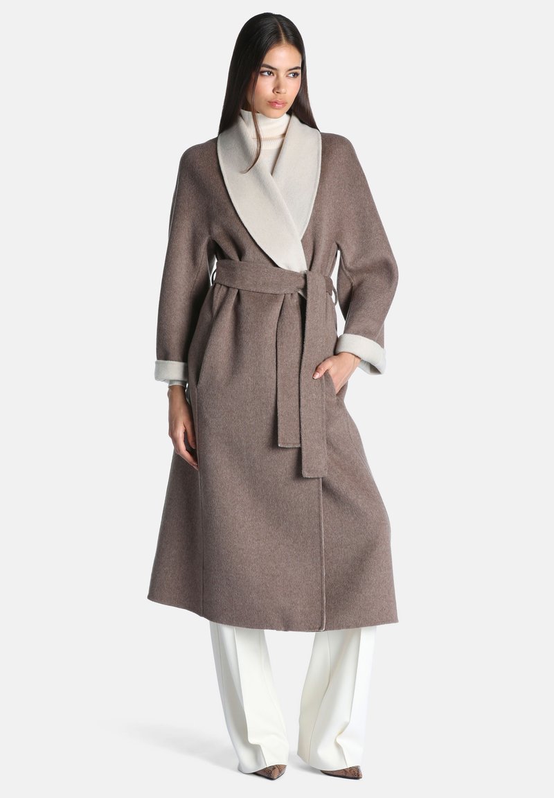 Femme portant un long manteau marron ceinturé avec un large col clair, un col roulé blanc, un pantalon blanc et des chaussures marron pointues, debout devant un fond uni.