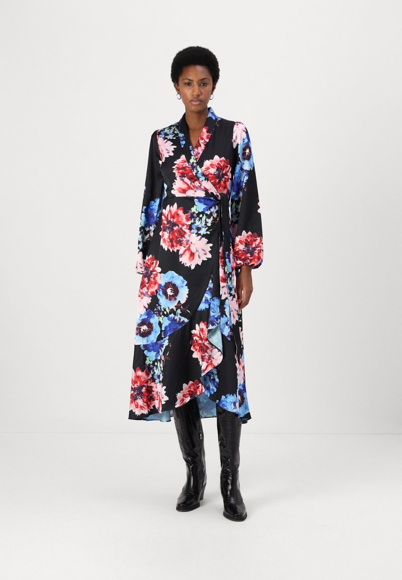 Robe portefeuille à fleurs avec un fond noir et des fleurs rouges et bleues, manches longues et un ourlet volé. Portée avec des bottes noires à mi-mollet.